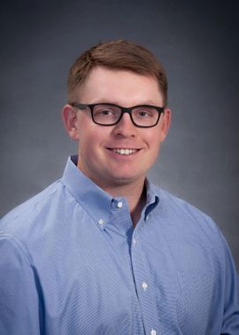 A professional headshot of a person with short brown hair and glasses, wearing a light blue shirt against a gray backdrop.