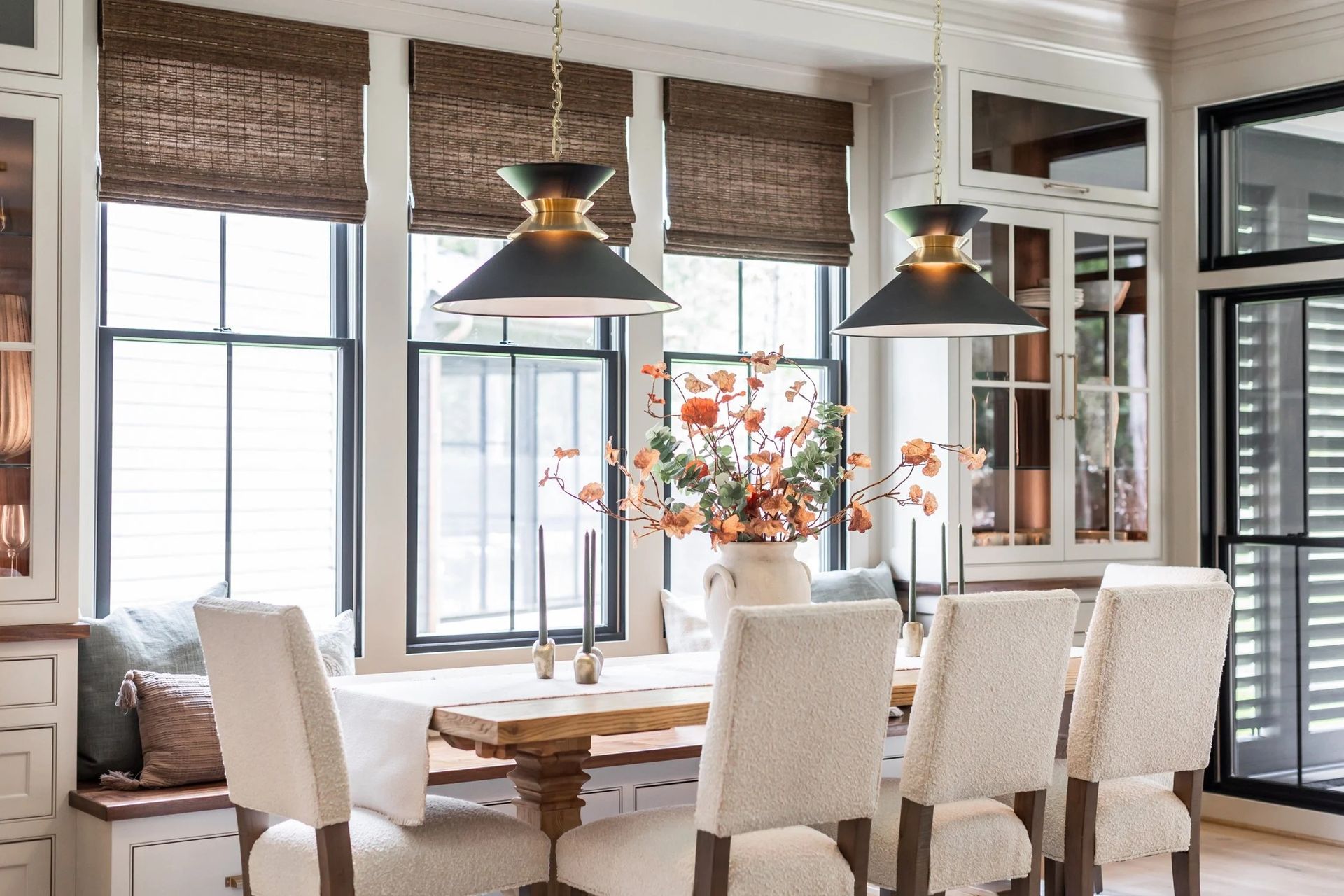 A bright, neutral dining area with a rustic wooden table, upholstered chairs, a window seat, and two modern pendant lights.