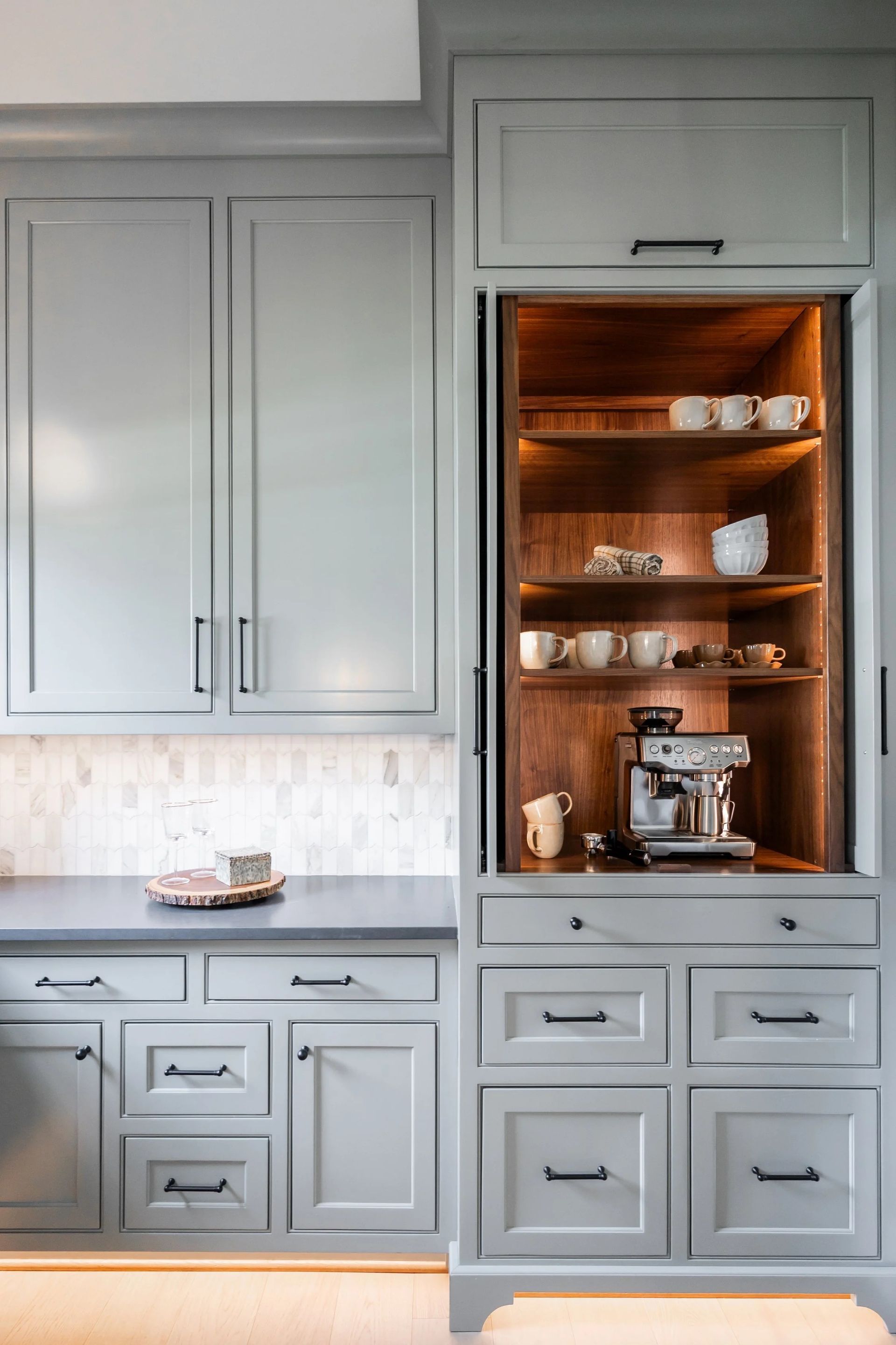 Gray kitchen cabinets with a built-in coffee station featuring a sliding door, wooden interior, and illuminated shelves.