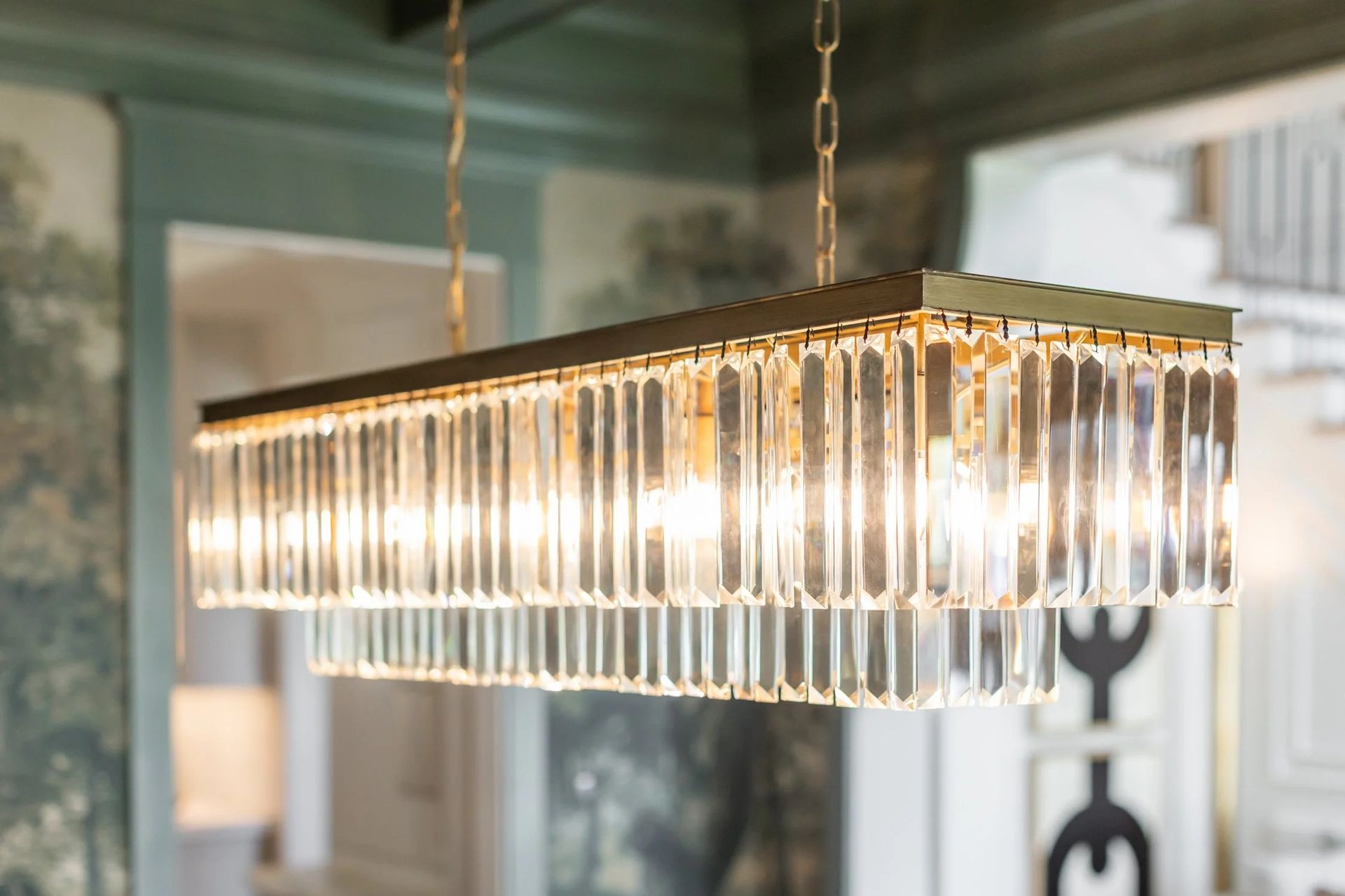 A rectangular, tiered crystal chandelier with a metallic frame suspended by chains in a room with patterned wallpaper.