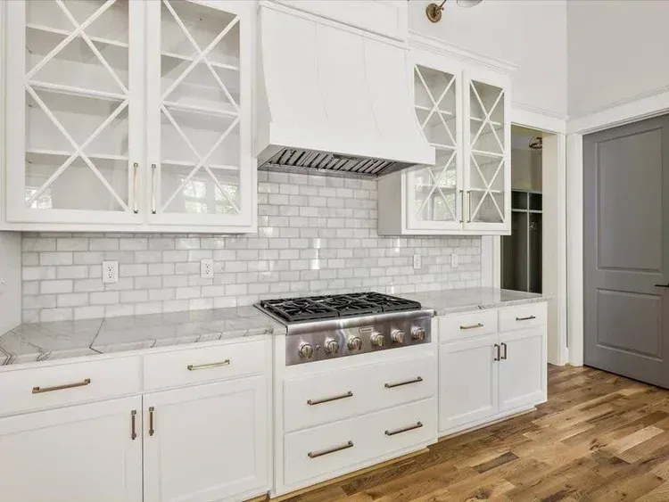 Bright white kitchen with glass-front cabinets, marble backsplash, stainless range, and wood floor