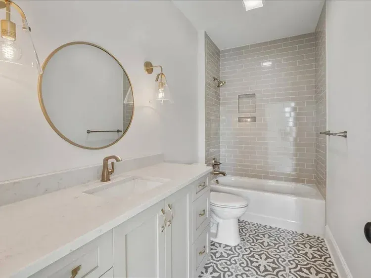 Bright white bathroom with vanity, round mirror, toilet, tub, and patterned black-and-white floor tiles