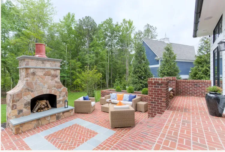 Brick patio with stone fireplace, wicker chairs, and brick seating area overlooking trees