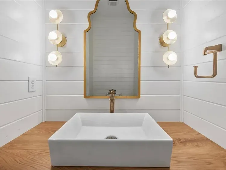 Modern bathroom vanity with rectangular white sink, gold-trimmed mirror, and wall sconces over wood countertop