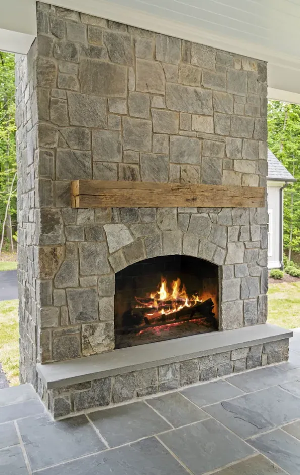 Stone outdoor fireplace with a burning fire and rustic wood mantel on a covered patio