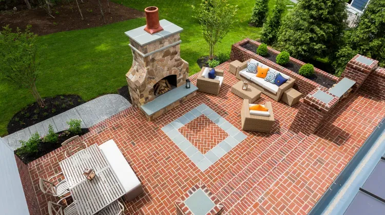 Brick patio with outdoor fireplace, cushioned seating, and a dining table beside a green lawn.