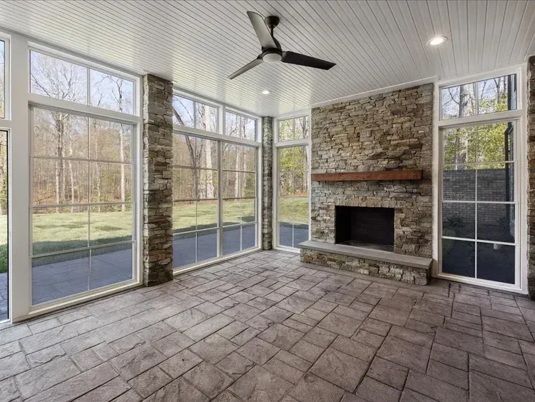 Bright sunroom with stone fireplace, tiled floor, ceiling fan, and tall windows overlooking a grassy yard