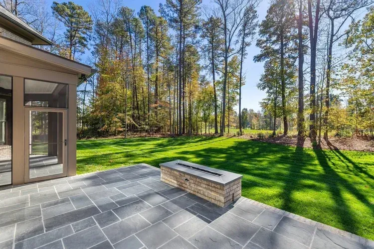 Patio with stone pavers and fire pit overlooking a sunny green lawn and wooded yard