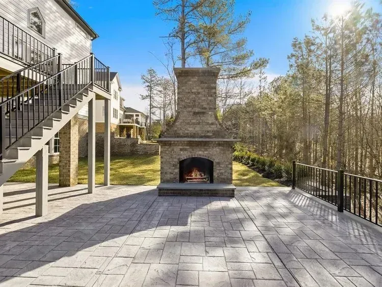 Stone outdoor fireplace on a paved patio beside a house, with stairs and trees in sunlight