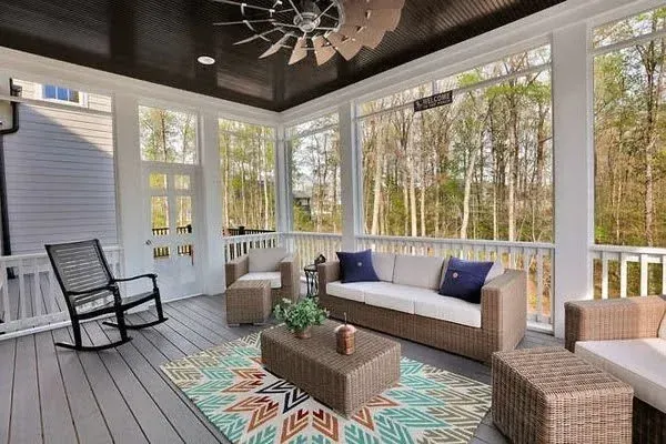 Covered porch with wicker seating, patterned rug, and large windows overlooking trees