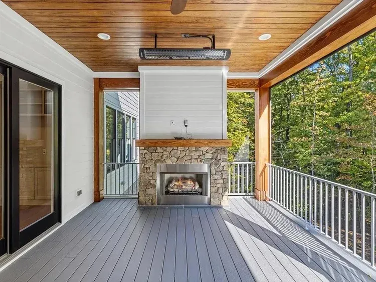 Covered porch with gray flooring, wood ceiling, stone fireplace, and white railing overlooking trees