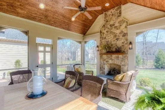 Bright sunroom with wicker seating, wood ceiling, stone fireplace, and large windows overlooking a grassy yard