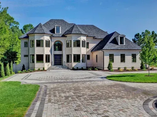 Large modern stone mansion with a curved driveway and landscaped front yard under a clear blue sky