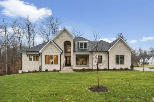 Large beige suburban house with a dark roof and arched entry, fronted by a wide green lawn and bare trees.