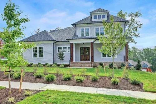 White two-story suburban house with a front porch, landscaped yard, and young trees under a blue sky