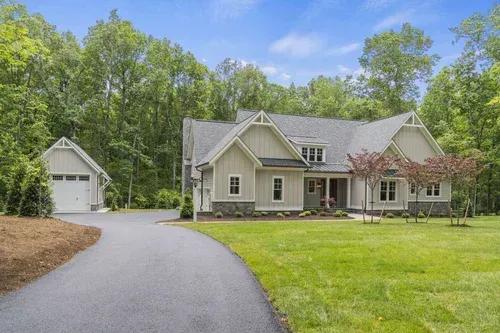 Single-story beige house with a detached garage, curved driveway, and green lawn surrounded by trees.