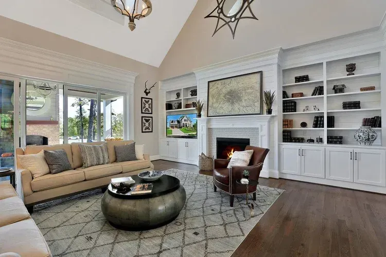 Bright living room with beige sofa, dark ottoman, fireplace, built-in shelves, and large windows.