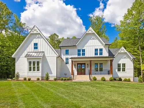 White farmhouse with gray roof, blue shutters, and a wide front porch on a green lawn under a bright sky