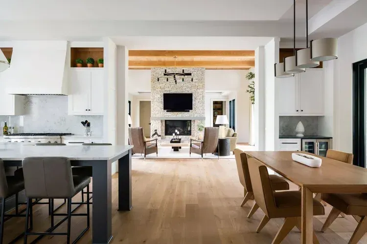 Modern open-plan kitchen and dining room with wood floors, white cabinets, and a fireplace in the background