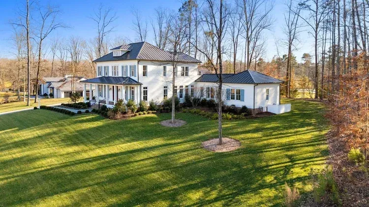 White two-story house with porch and attached garage on a large green lawn, surrounded by bare trees.
