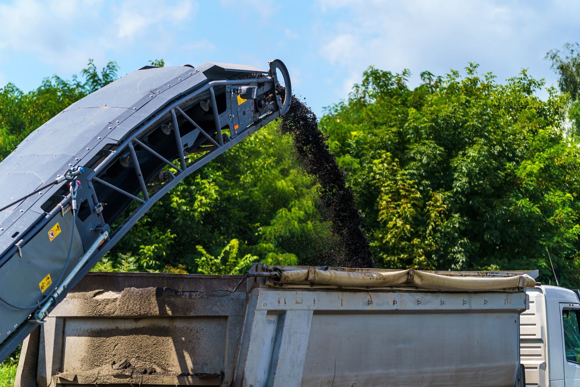 Heavy machinery is pouring material on a job site. Blue sky and green foliage are in the background.