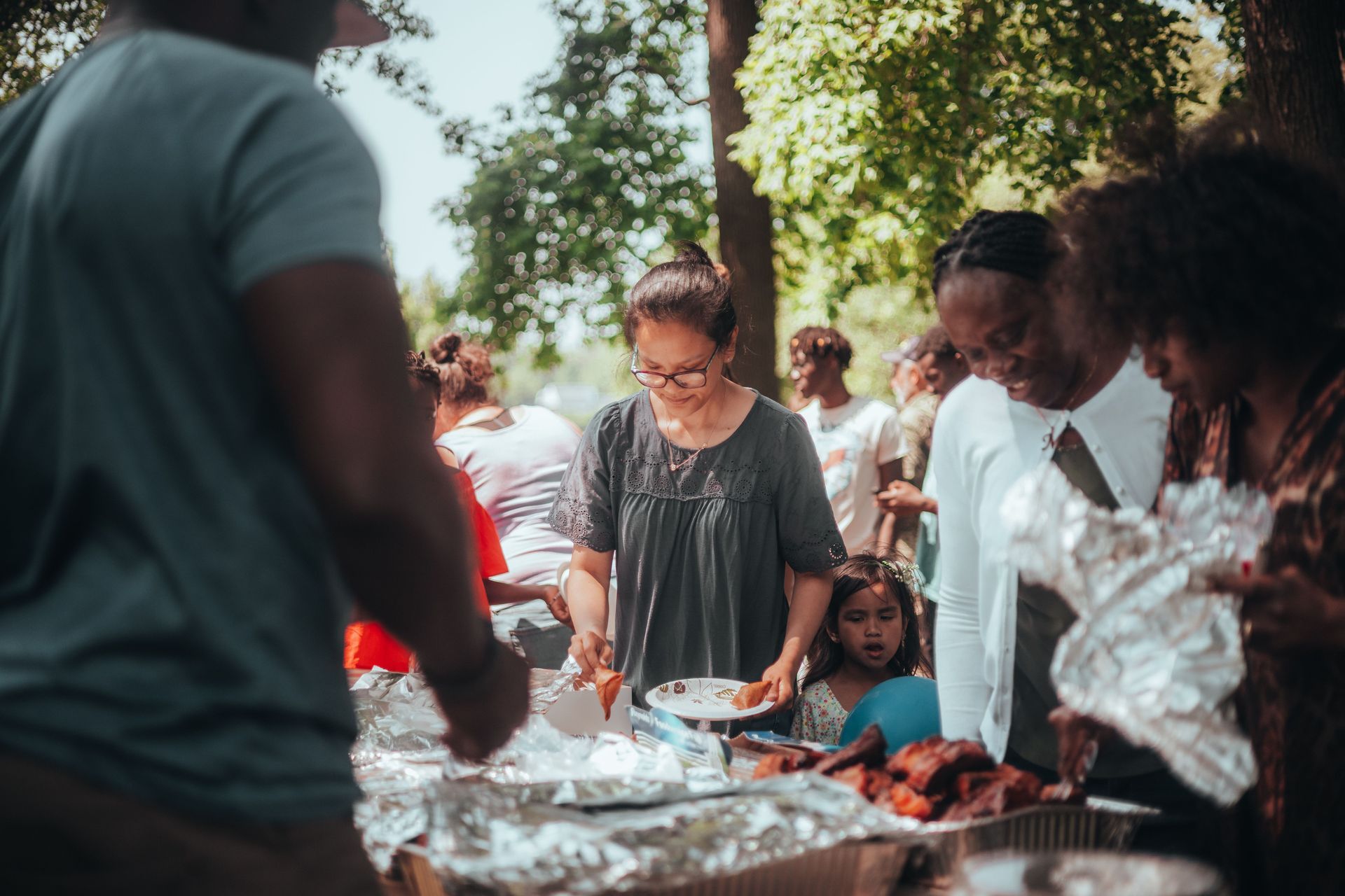 People serving food at an outdoor gathering under trees, with trays on a table and seated guests nearby