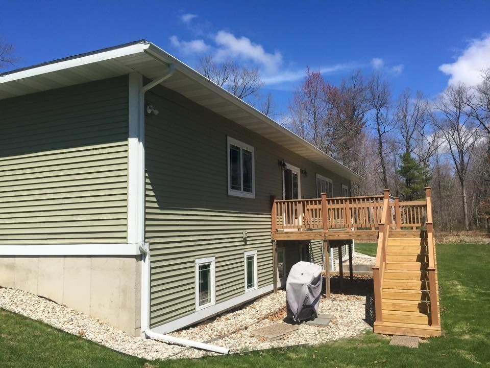 Green-sided house with a wooden deck and stairs, set in a grassy yard under a bright blue sky.