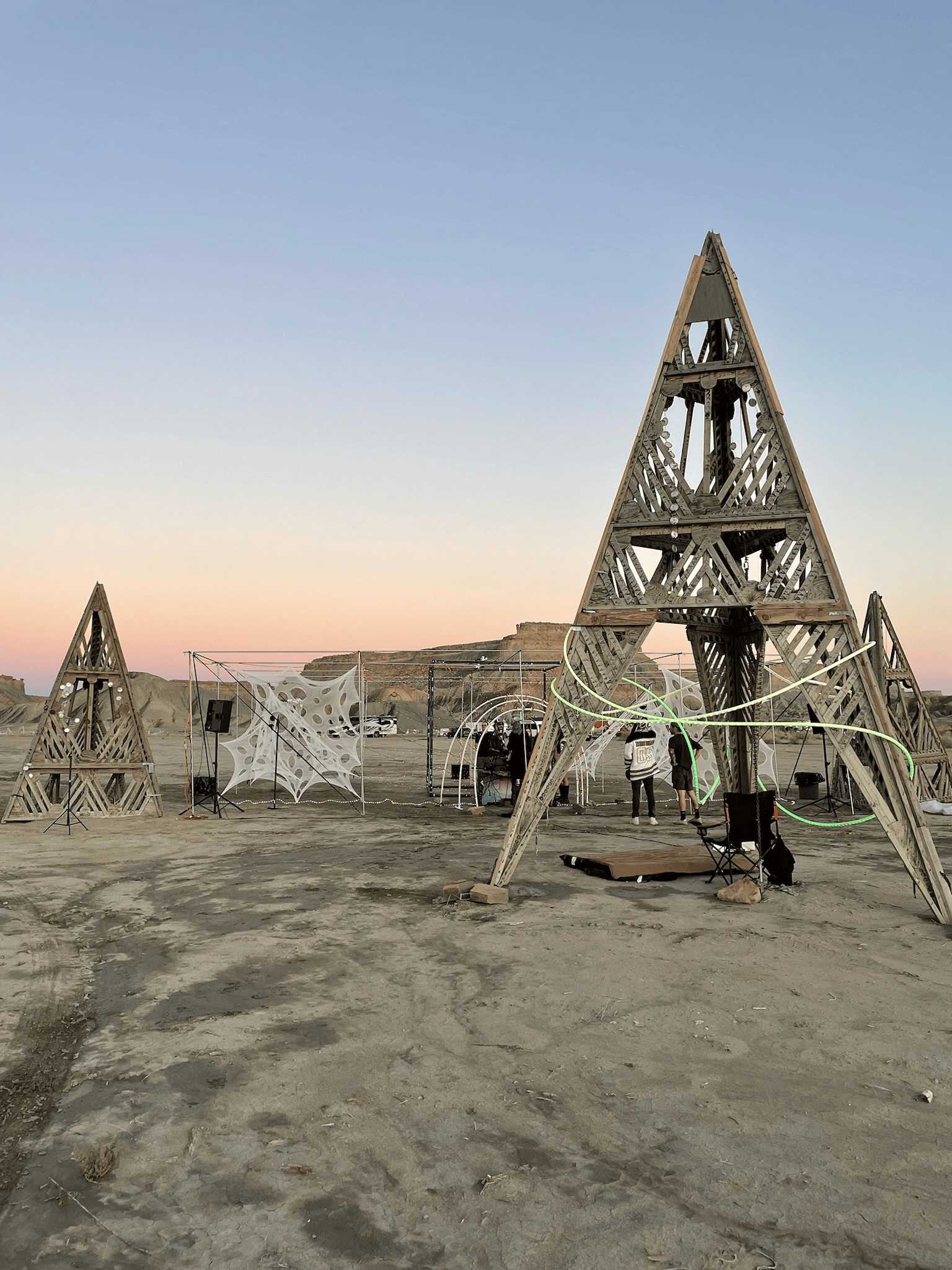 Wooden triangular structures on a sandy expanse, with people and city backdrop under a dusk sky.