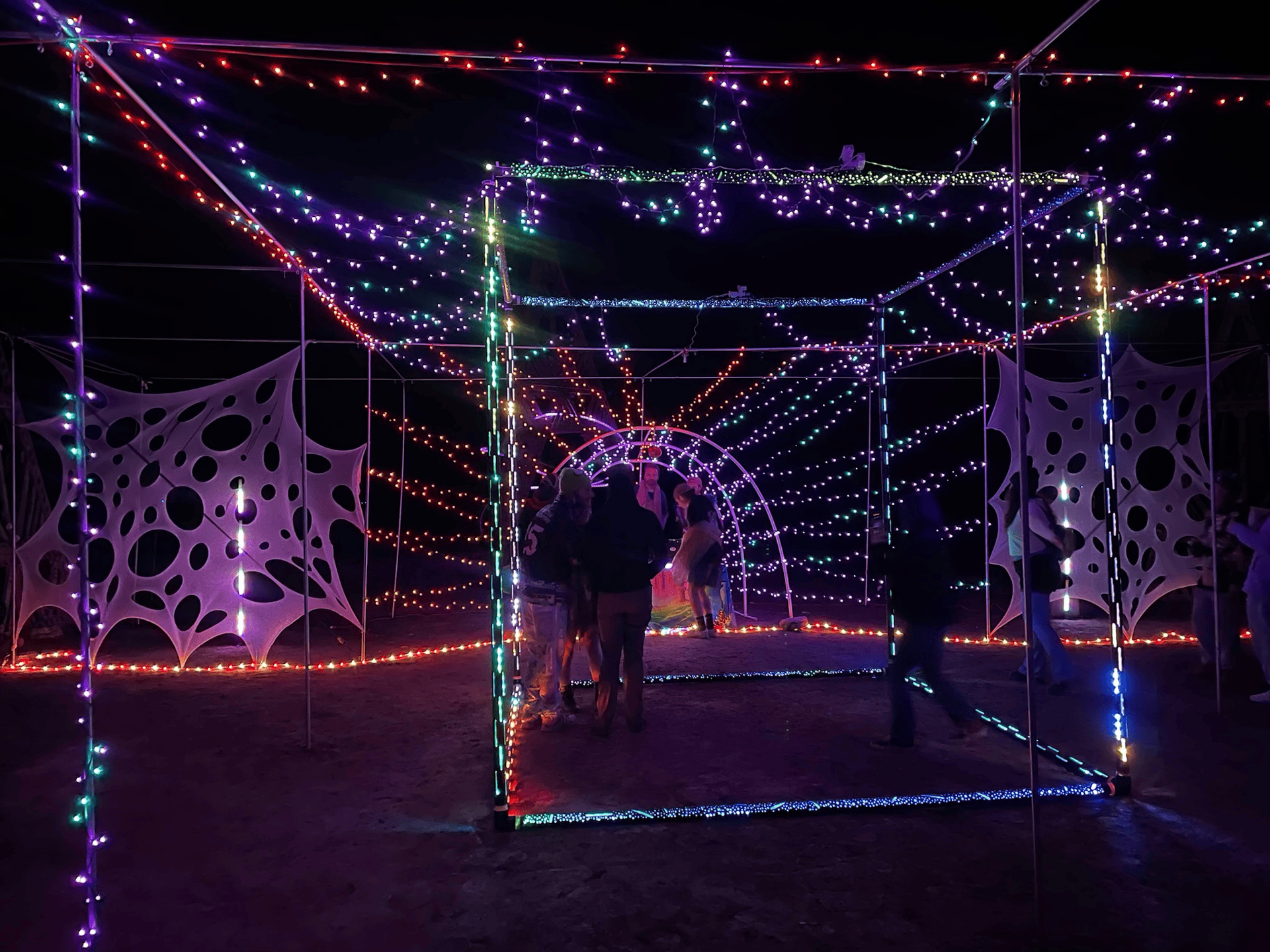 Illuminated cube-shaped tunnel at night, strung with colorful lights. People stand inside.