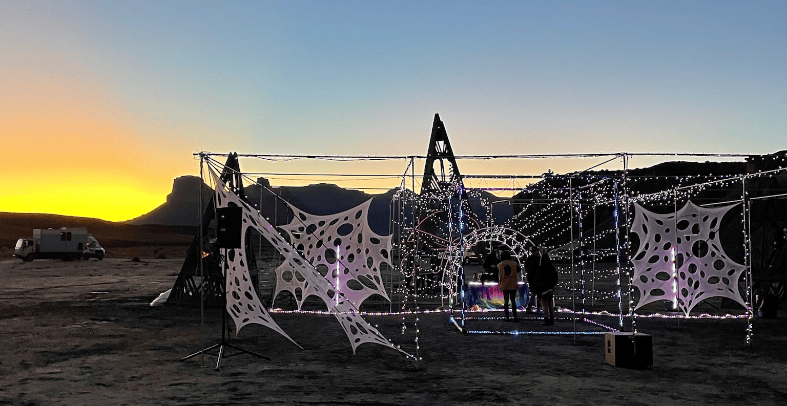 Outdoor art installation with geometric shapes and lights against a sunset. People stand nearby.