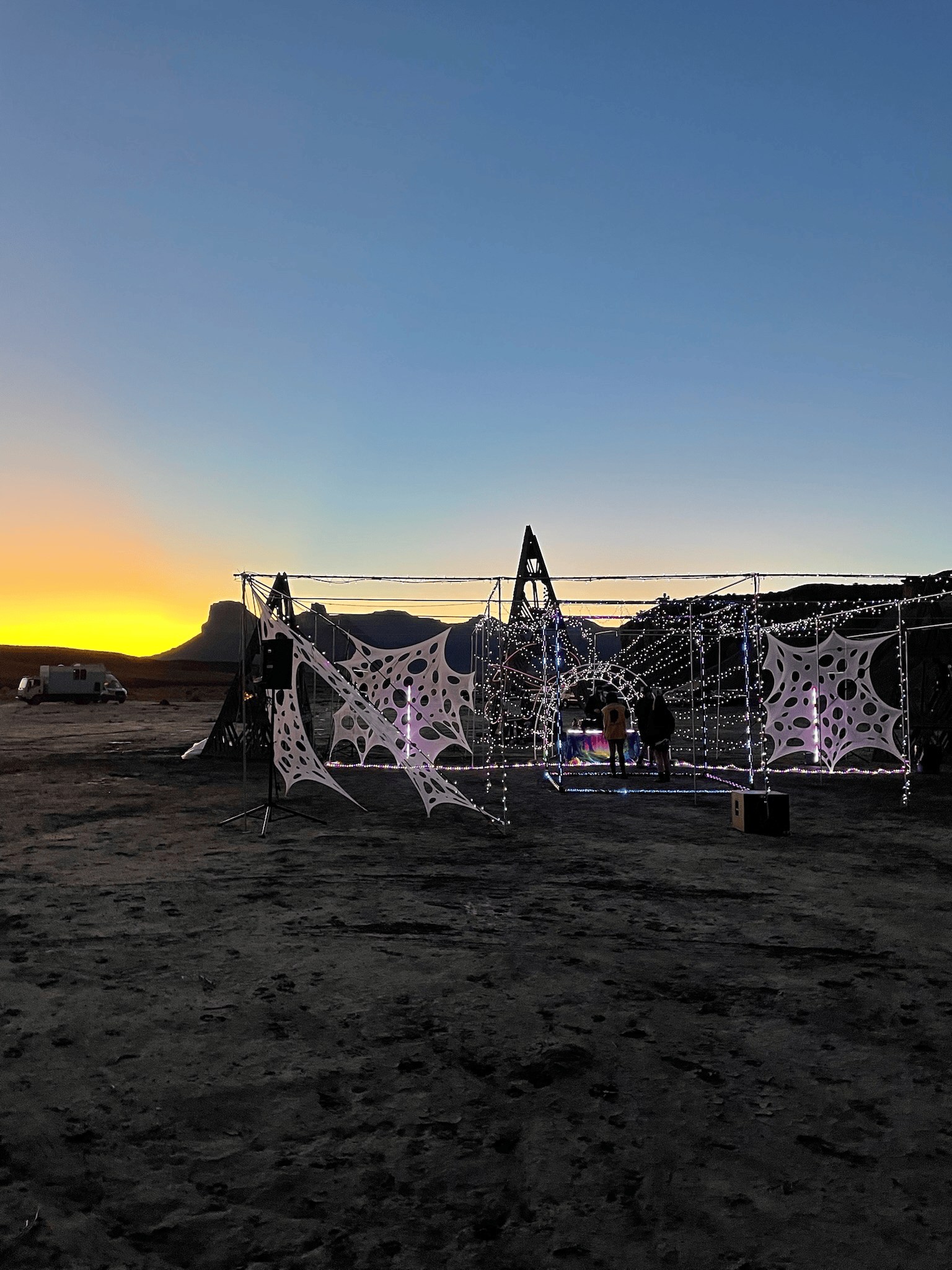 A desert landscape at dusk with a geometric art installation illuminated in white. A camper is visible in the distance.