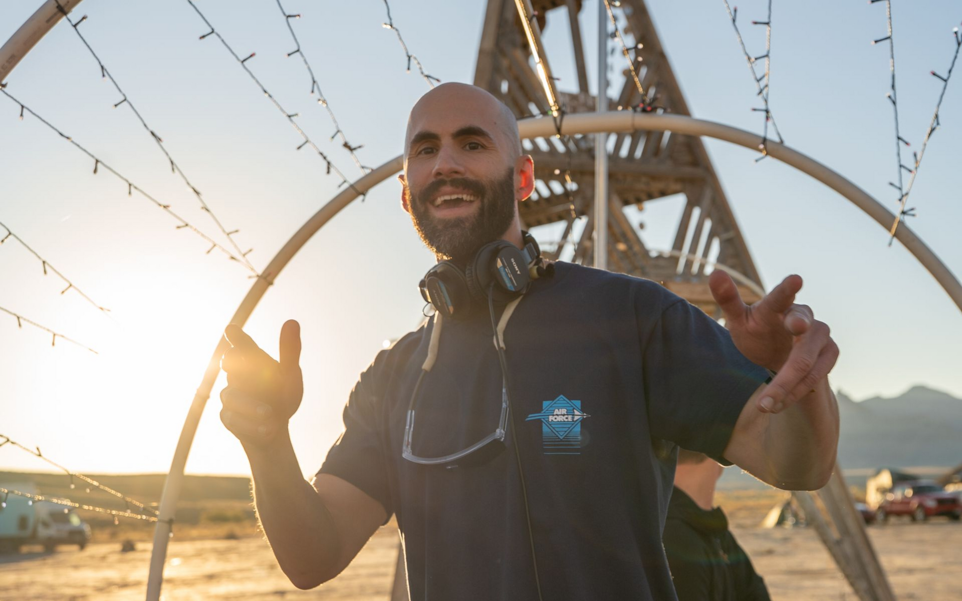Man with headset and beard smiles, gesturing.  Sunset, open desert, and structure with lights in background.