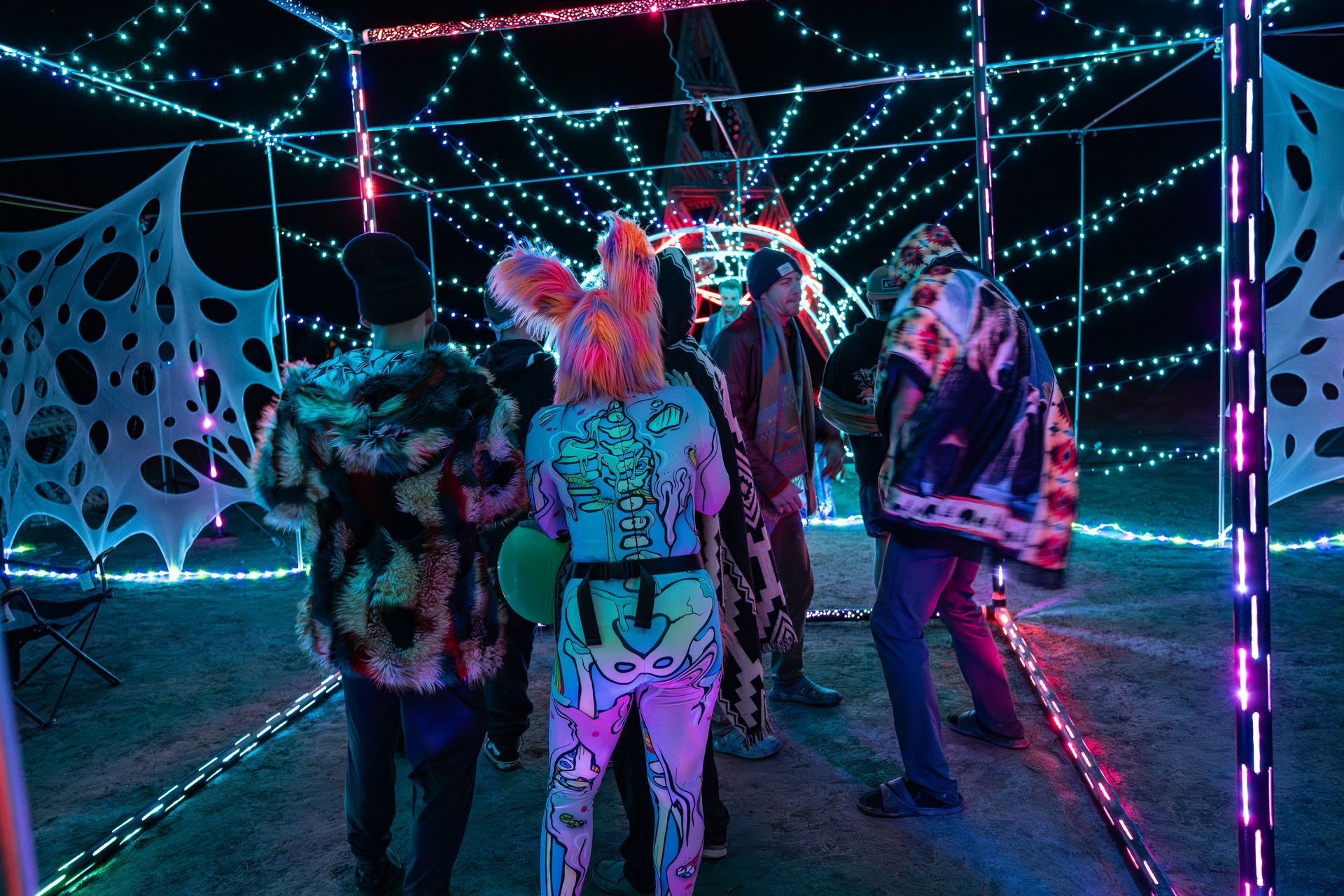 People inside a glowing geometric structure decorated with lights and webs. One wears a bunny mask and body paint.