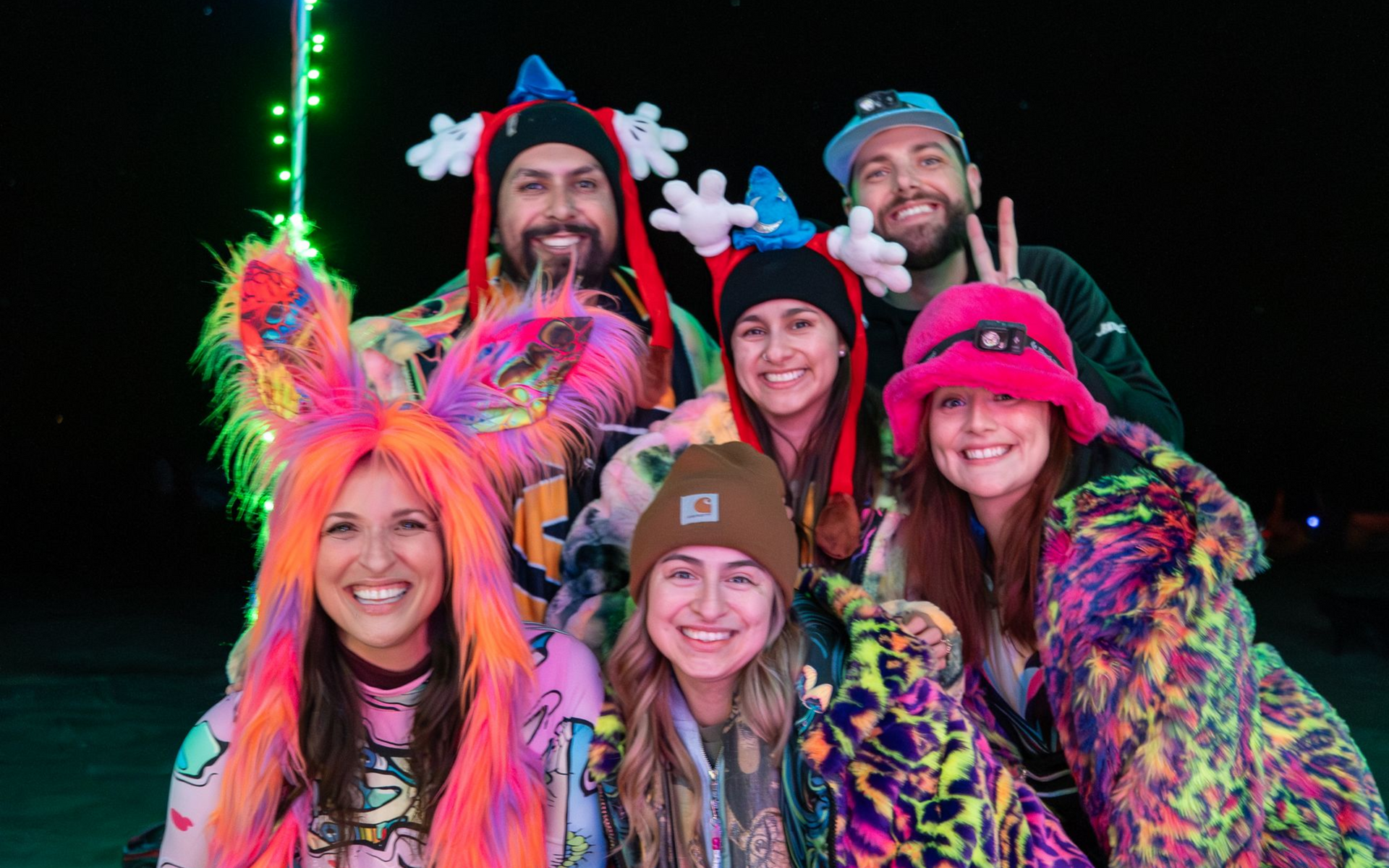 Group of six people smiling, wearing colorful fur costumes and hats, illuminated by green lights.