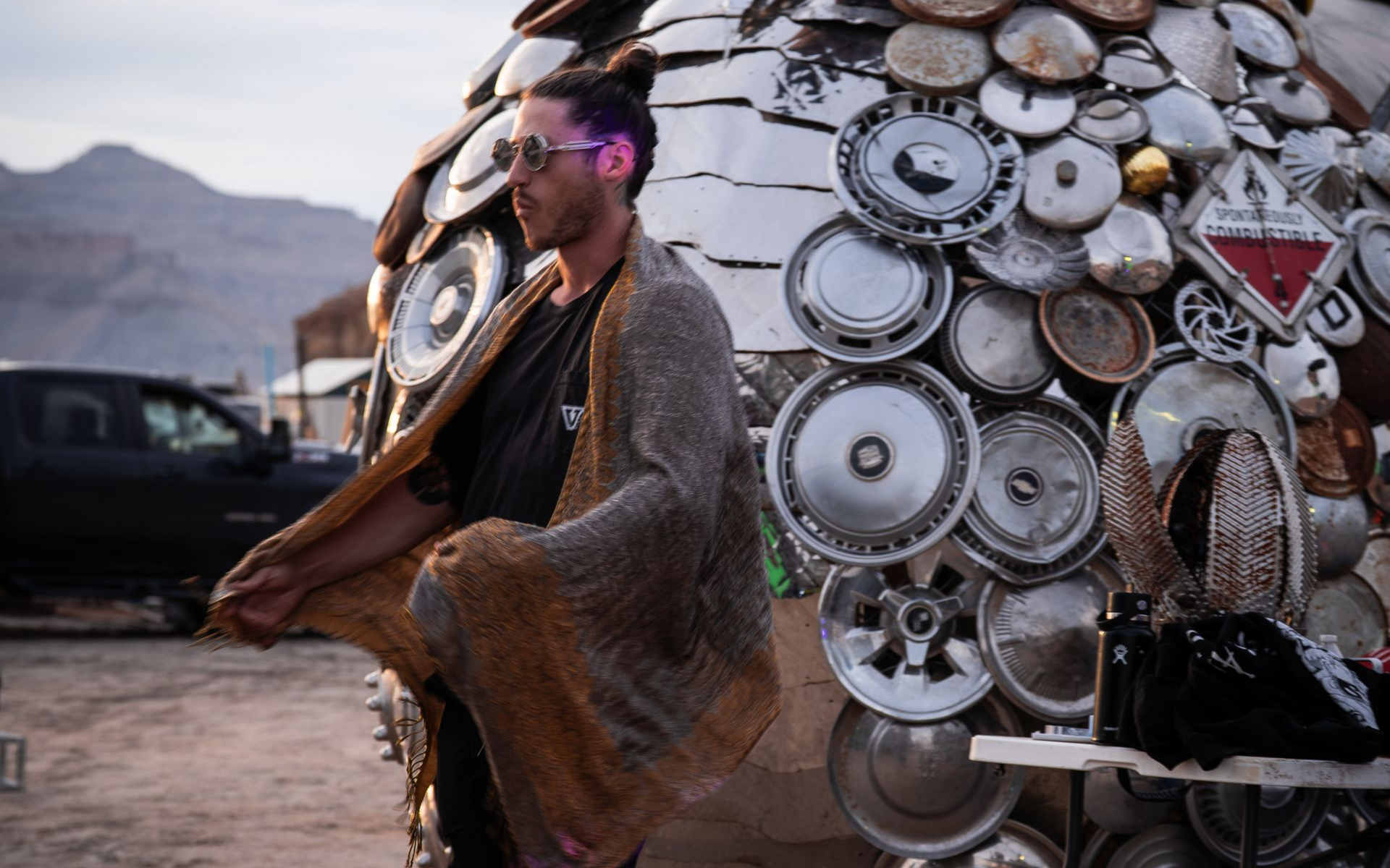Man in metallic cape by a sculpture made of hubcaps, desert setting.