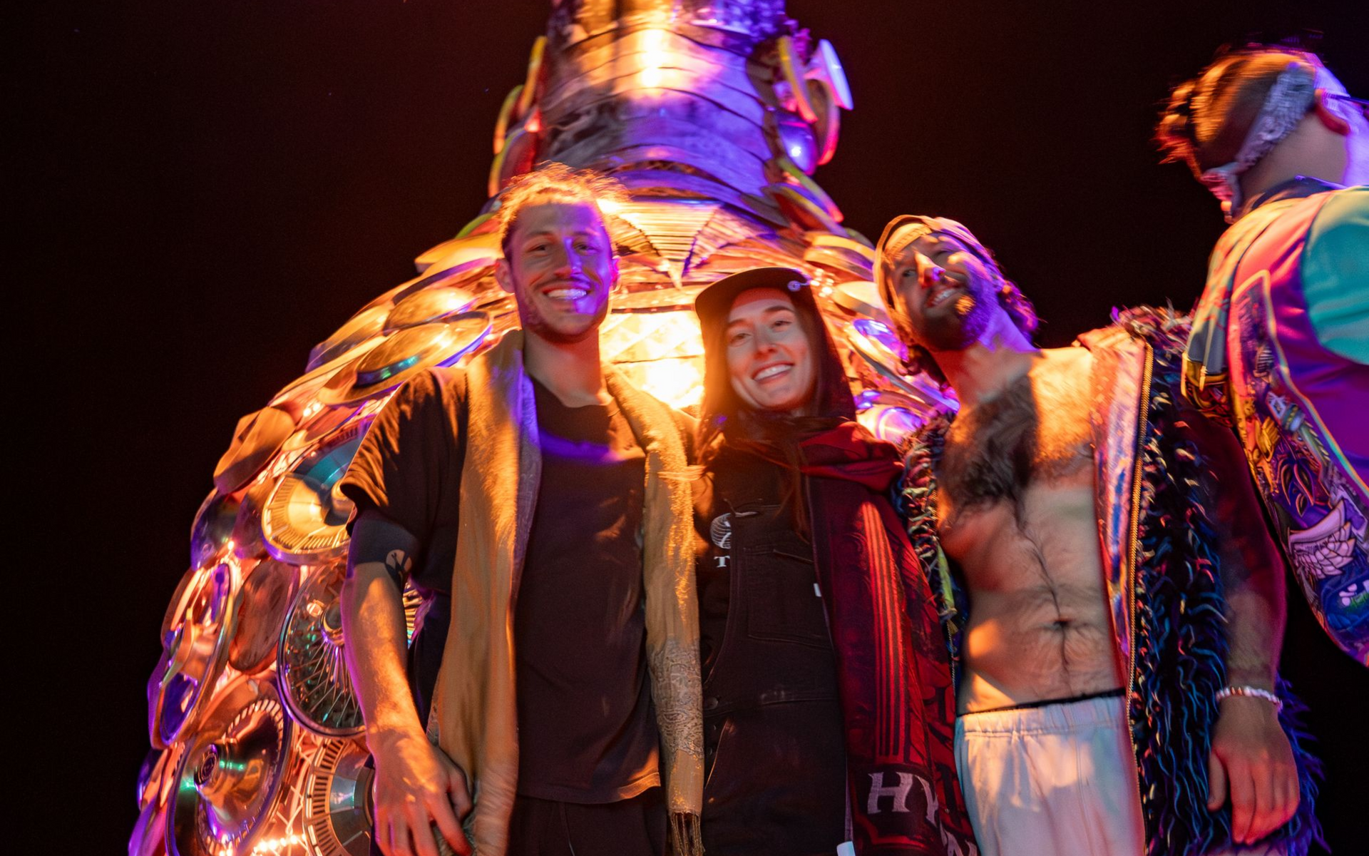 Three people pose in front of a colorful sculpture. Dark setting. Smiles, open-air clothing, and headwear.