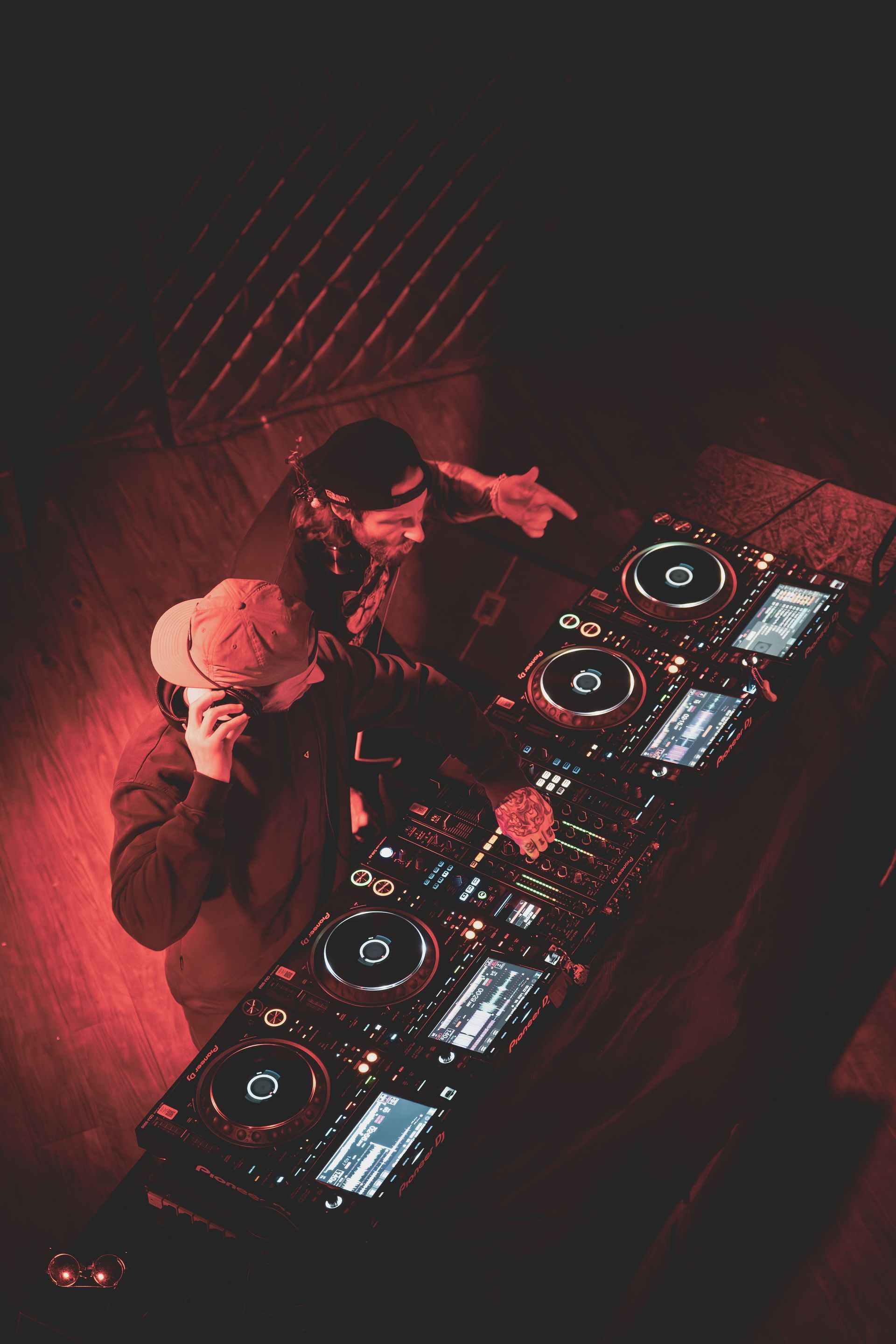 Two DJs in a dimly lit club playing music. Red lighting illuminates the equipment and their faces.