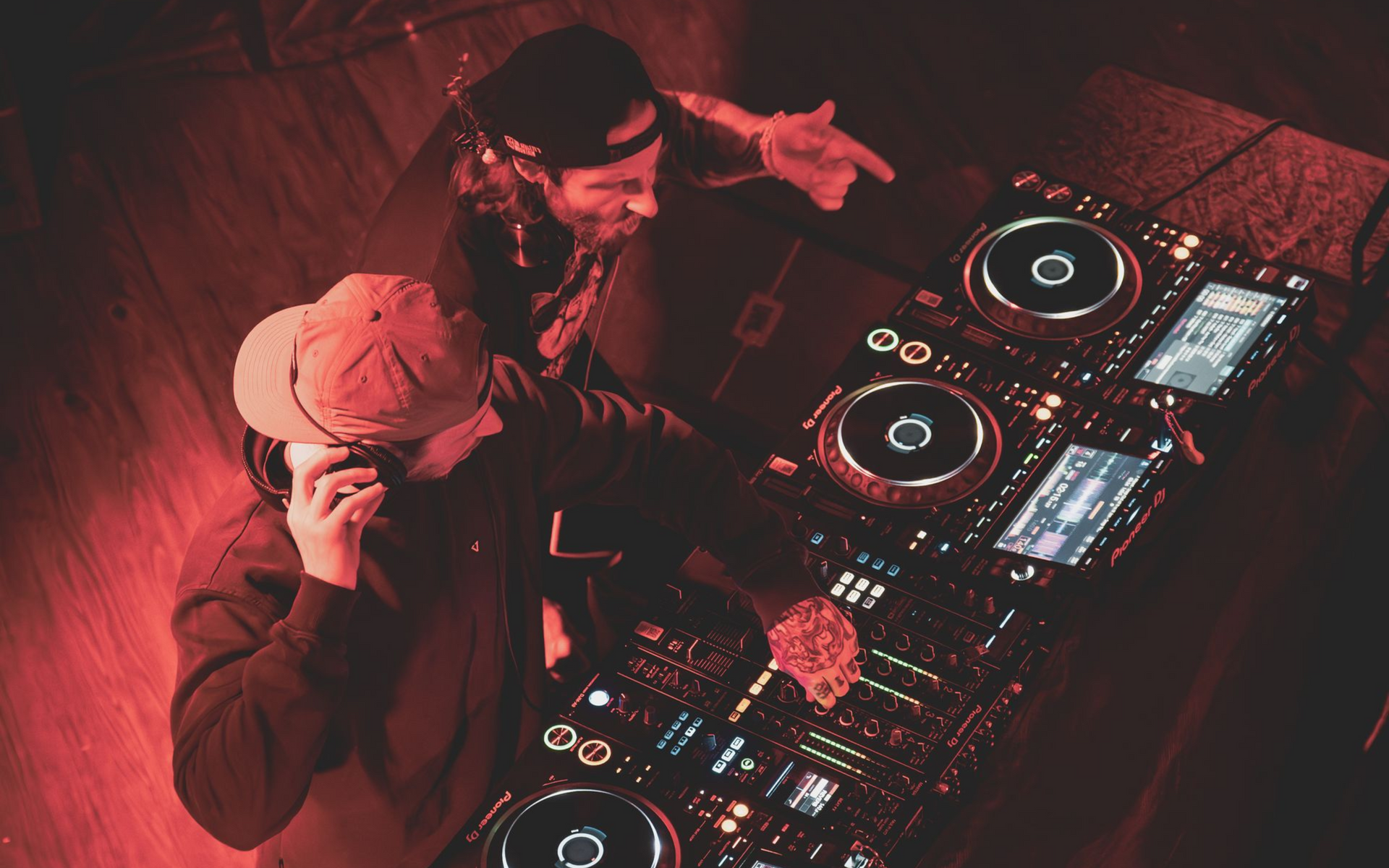 Two DJs operating a mixing console in a dimly lit setting, bathed in red light. One points, the other adjusts settings.