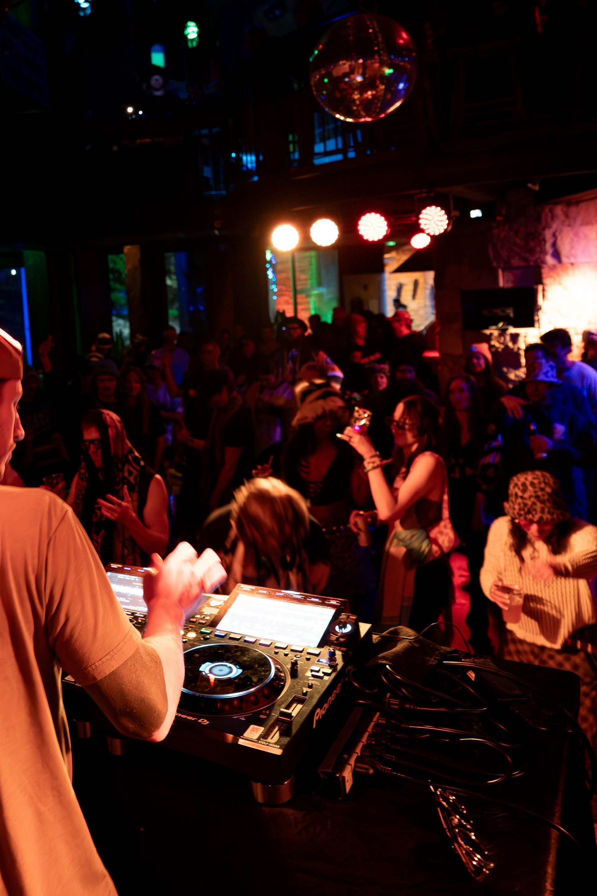 DJ at a crowded nightclub, playing music, audience dancing under disco ball and colored lights.