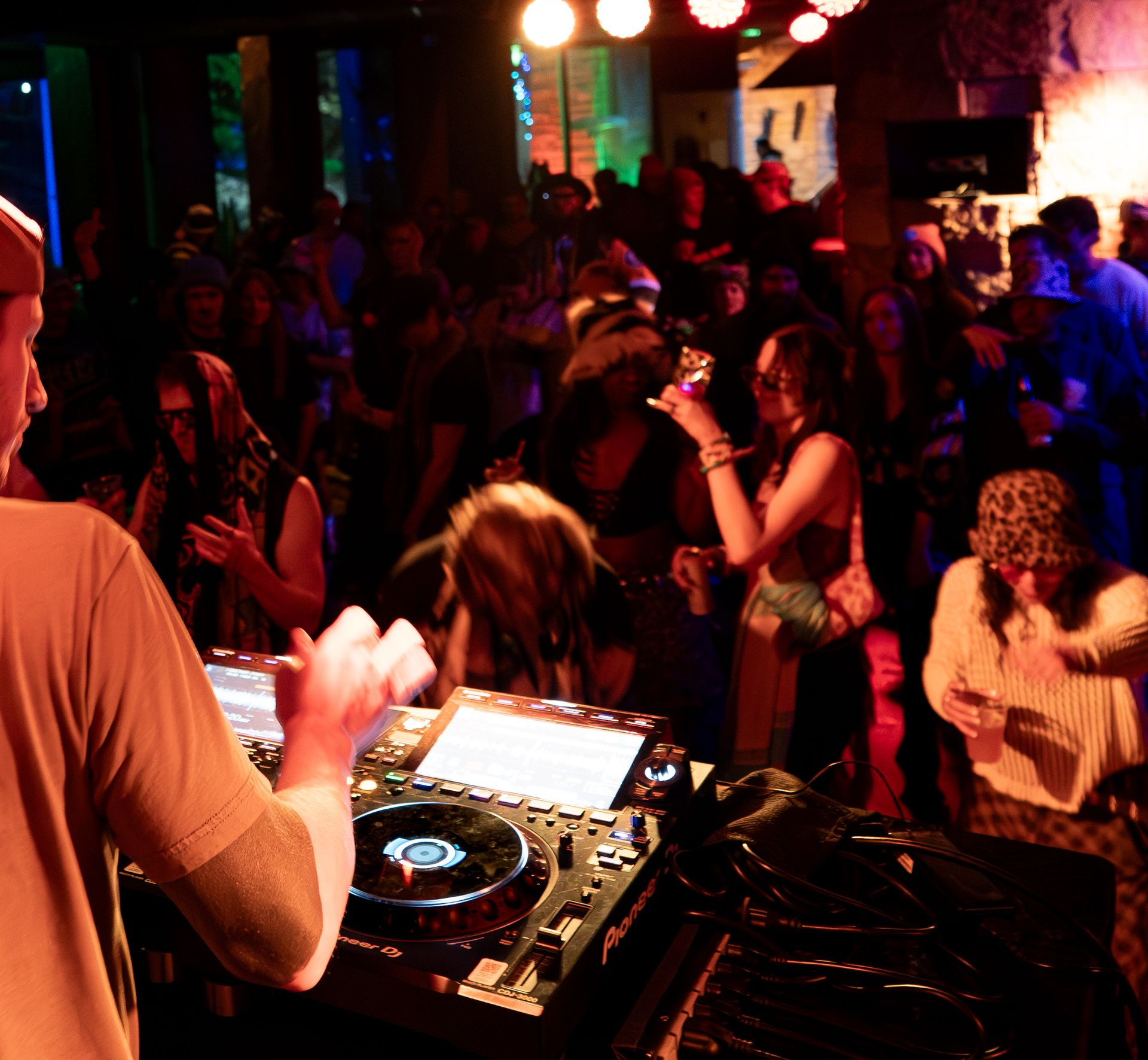 DJ performing at a crowded club with people dancing, colorful lights, and a stone wall backdrop.