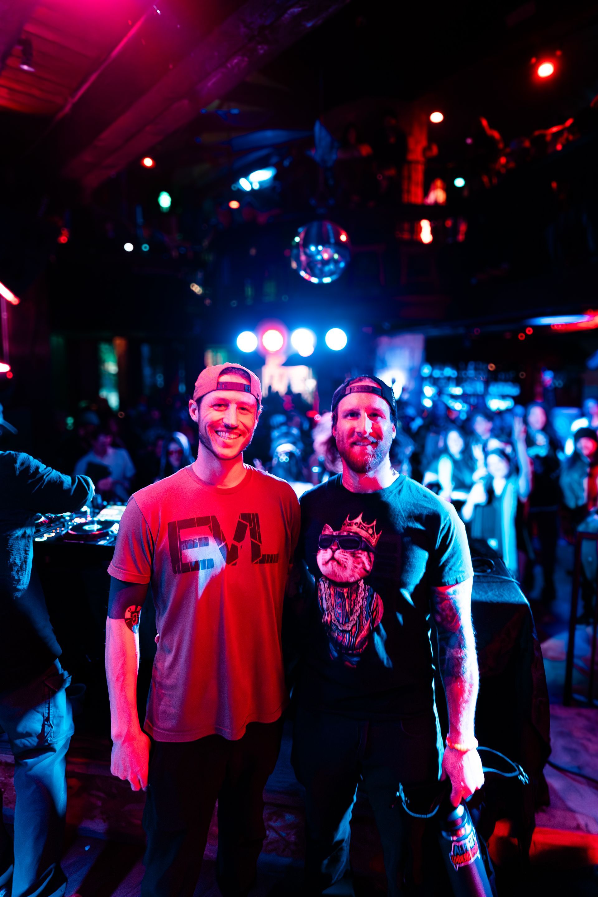 Two men posing at a concert, one in a red shirt, the other with tattoos, both with baseball caps, standing in front of a crowd.