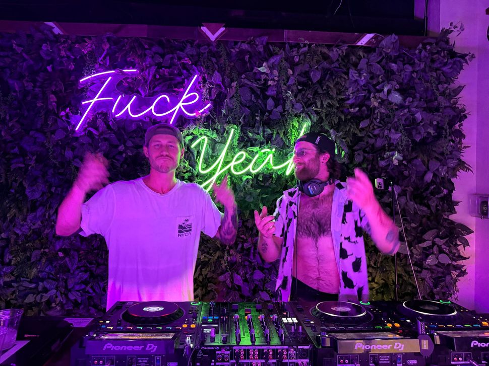 Two DJs at a party, gesturing enthusiastically in front of a neon sign that says 
