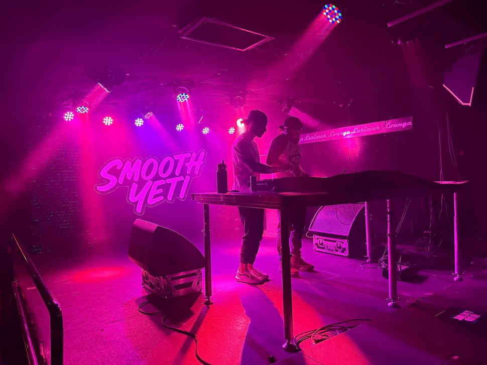 Two people DJing at a club, lit by pink and purple stage lights. The sign behind them says 
