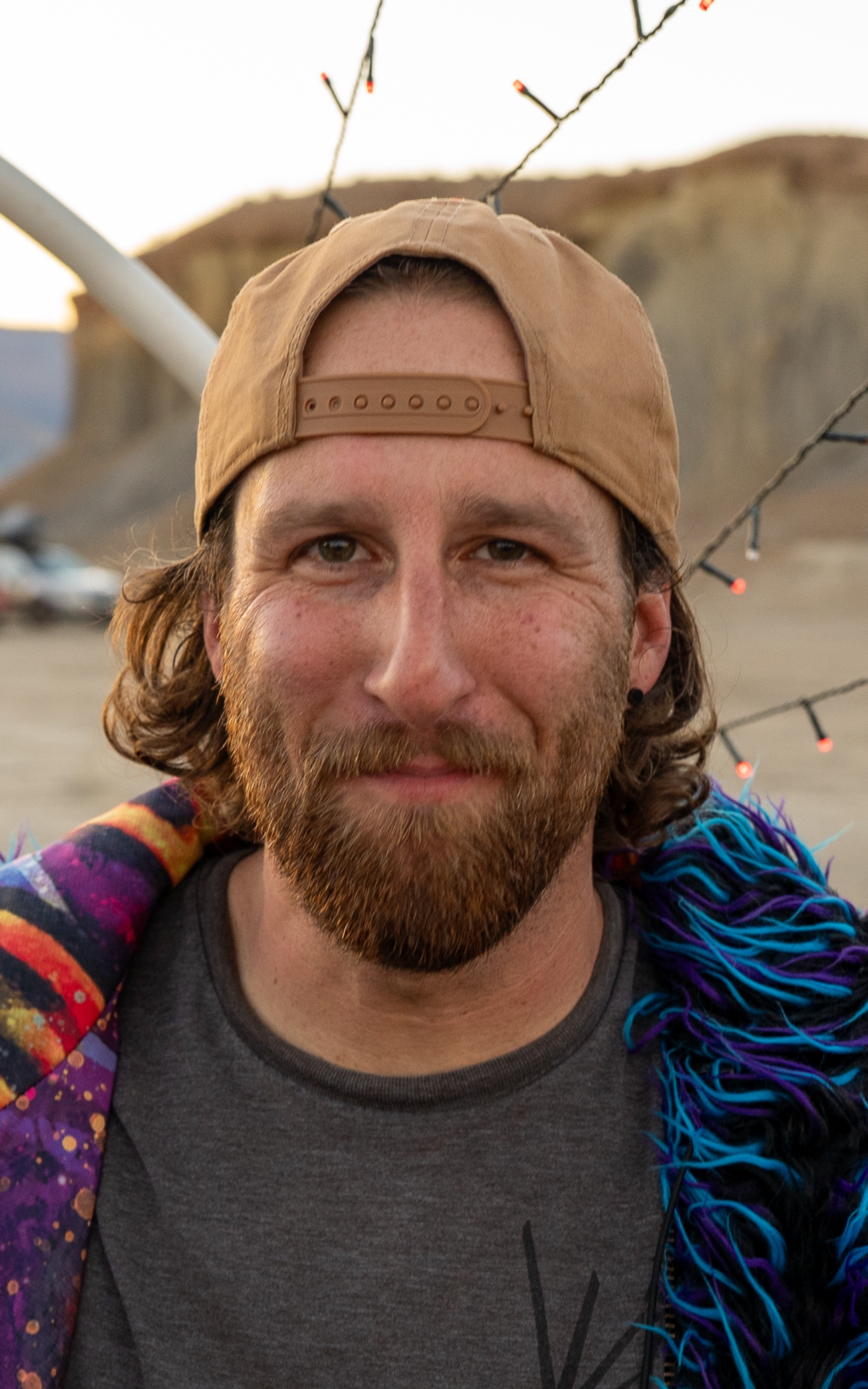 Man with brown cap and beard smiling, wearing a colorful, furry garment, outdoors.
