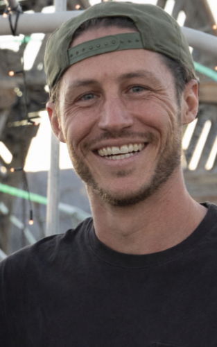 Man wearing a green cap smiles widely; a slight beard and black shirt. Outdoor setting, warm lighting.
