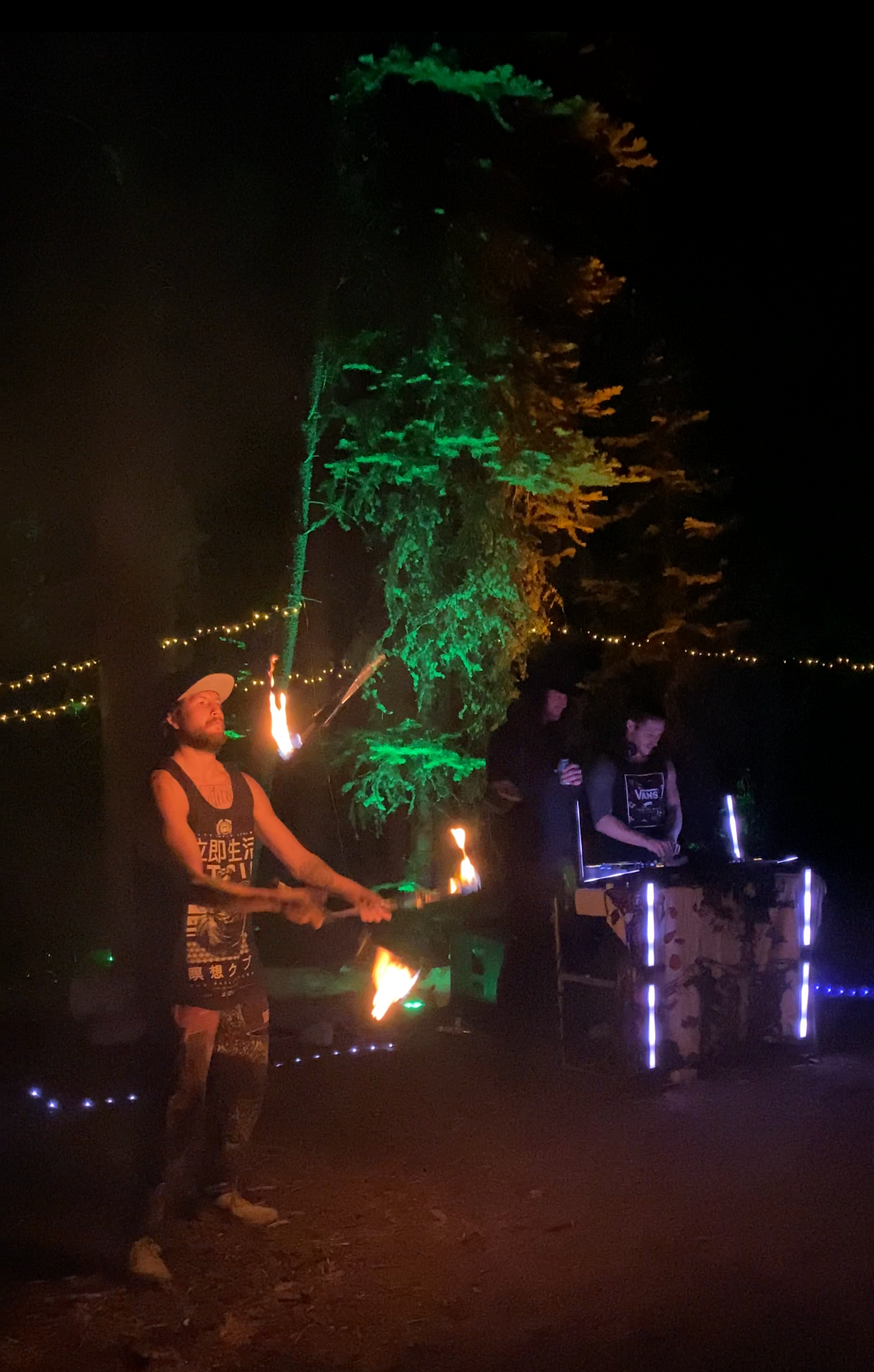 Person twirling fire stick near a tree lit in green, with DJ playing music and lights in a dark outdoor setting.