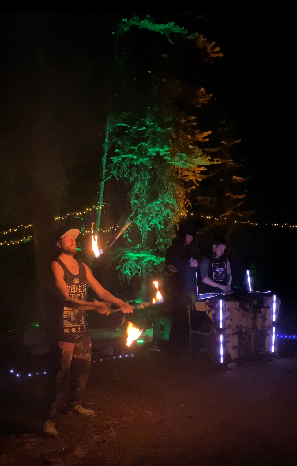 Person twirling fire stick near a tree lit in green, with DJ playing music and lights in a dark outdoor setting.