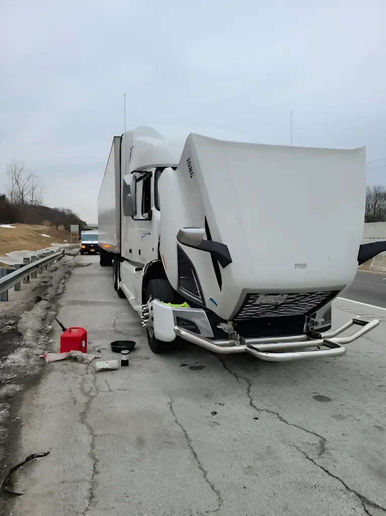White semi-truck with its hood open on a highway shoulder; red gas can and tools nearby. Cloudy sky.