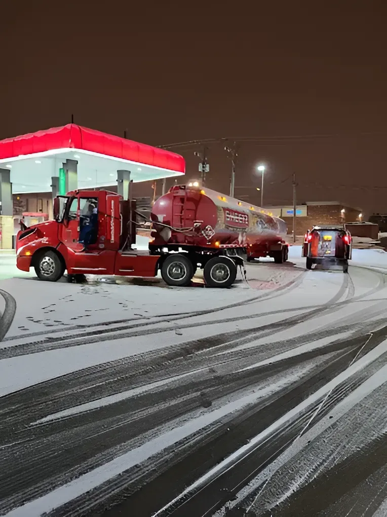Red fuel truck at a gas station at night, refilling tanks in snowy conditions.