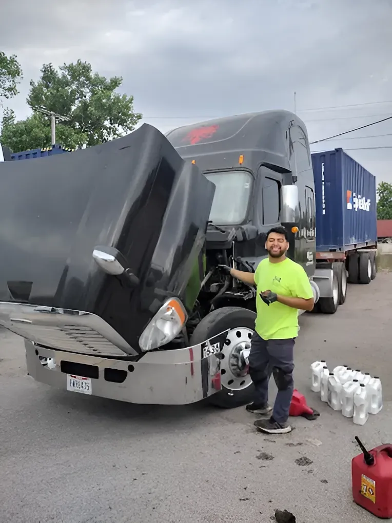 Man in a green shirt working on a black semi-truck with the hood open outside, containers on the back, sunny day.